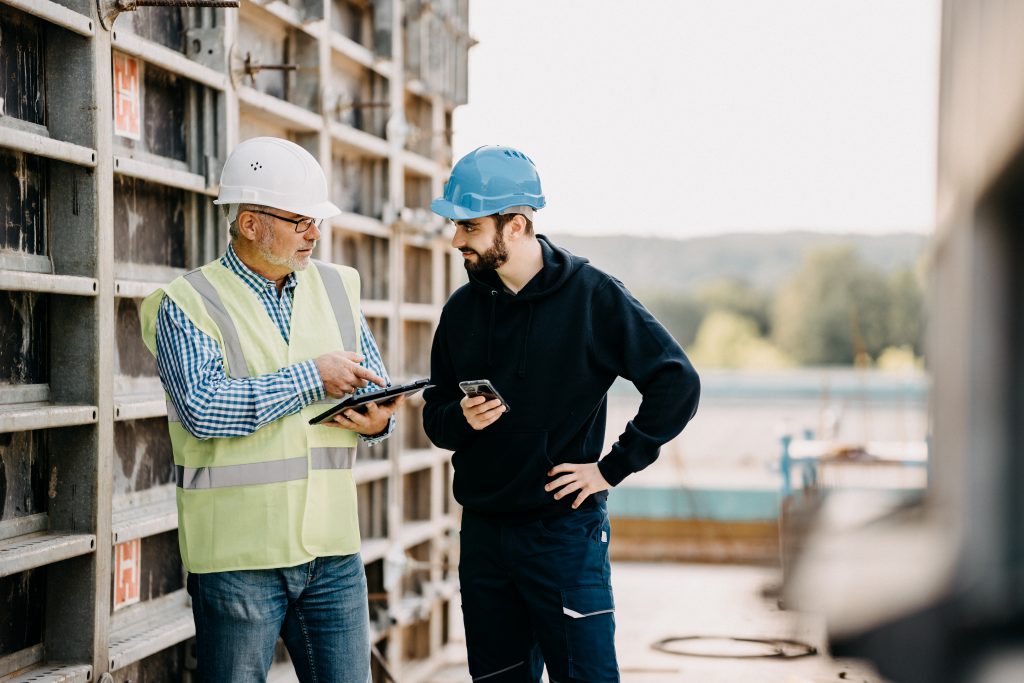 Zwei Mitarbeiter auf der Baustelle mit Helm und Tablet und Smartphone in der Hand