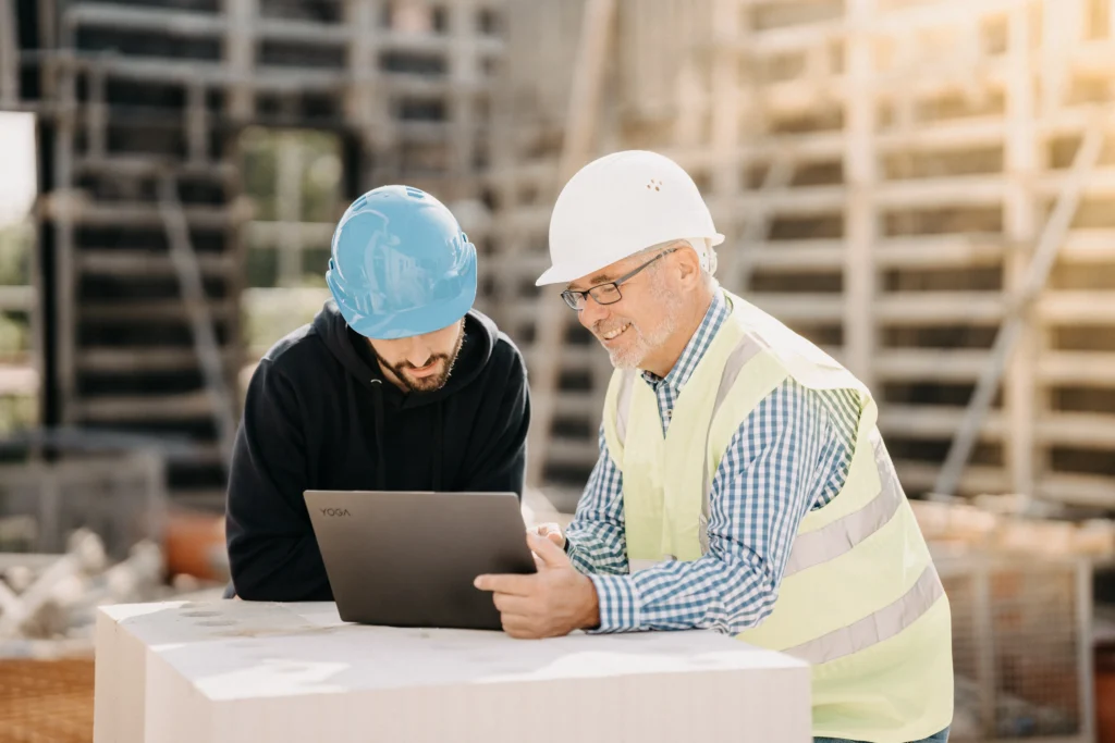 Zwei Bauarbeiter prüfen digitale Projektdaten auf einem Laptop auf der Baustelle und optimieren Abläufe durch moderne Software, die auch eine präzise Zeiterfassung und Ressourcenplanung direkt vor Ort ermöglicht.