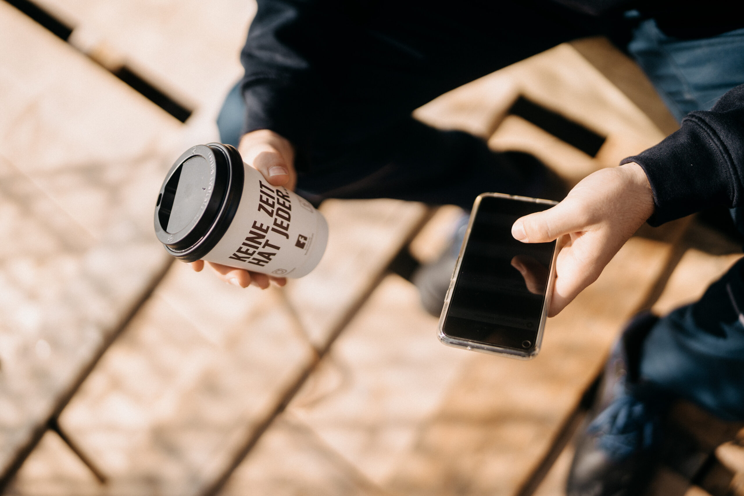 Handwerker hält Smartphone und Kaffeebecher mit „Keine Zeit hat jeder“ – mobile Zeiterfassung auf der Baustelle.