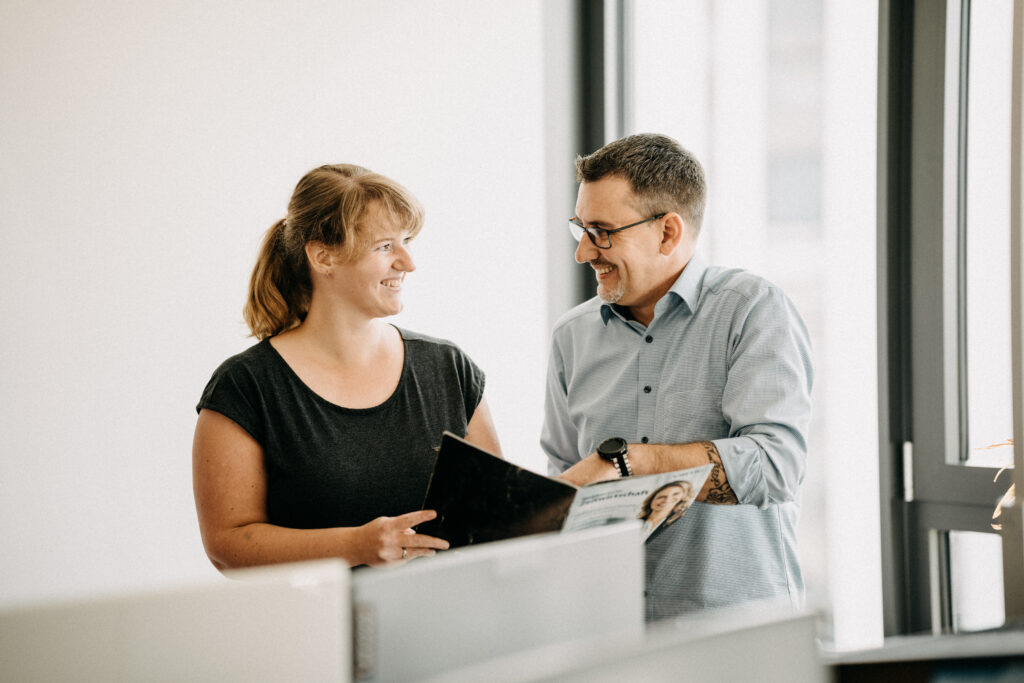 Zwei Mitarbeitende im Büro tauschen sich freundlich über Dokumente aus und besprechen Aufgaben rund um digitale Arbeitsprozesse.