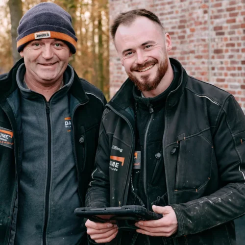 Zwei Handwerker stehen lächelnd auf der Baustelle und nutzen ein robustes Tablet für digitale Dokumentation und Zeiterfassung